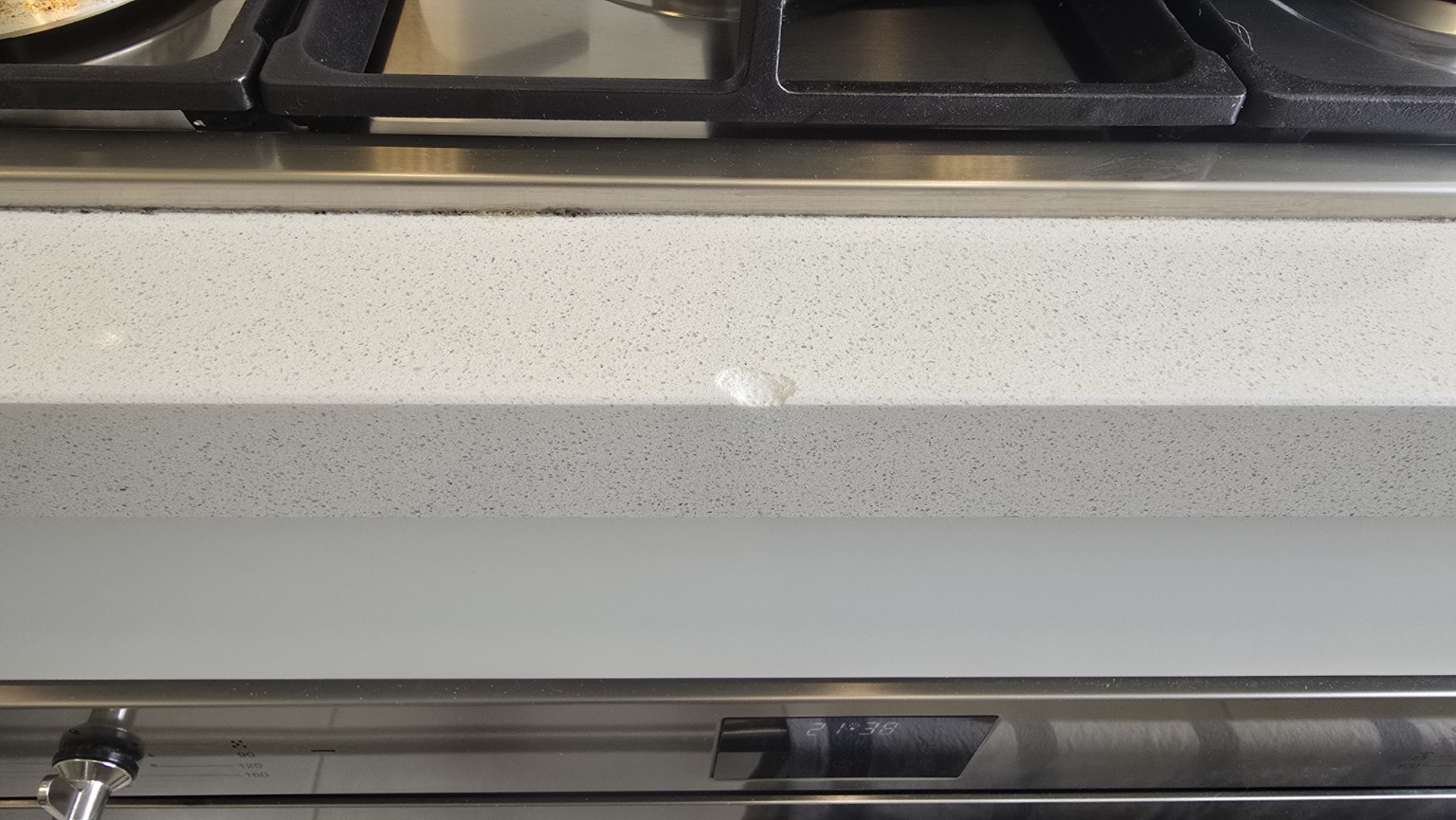 Chip on the top front edge of a pale speckled stone benchtop in front of a cooktop, before repair
